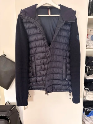 Mörkblå colmar cardigan  - Snygg mörkblå cardigan med stickade ärmar och huva. Jackan har quiltad front och huva i glansigt material, medan ärmarna är i grovstickad bomull. Två fickor med dragkedja framtill och dragsko i nederkant för justerbar passform.