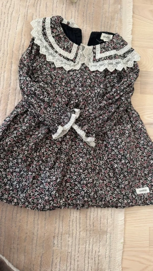 Blommig klänning med spets från Newbie - Supersöt blommig kortklänning från Newbie med vit spetskrage och rosett. Klänningen har små blommor i rosa, vit och grön på mörk botten. Perfekt för vår och sommar med sin romantiska look och fina detaljer.