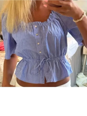 Blårandig blus med knyt och knappar - Söt blårandig blus med korta ärmar, knappar framtill och knyt i midjan som ger en fin form. Blusen har en lätt croppad passform och resår vid halsen. Perfekt till jeans eller kjol för en fräsch look.