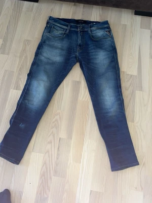 Blå slim fit jeans från Replay - Säljer ett par blå slim fit jeans från Replay med snygga slitningar och tvättade detaljer. Jeansen har klassisk femficksdesign, dragkedja och knapp i midjan samt tydliga kontrastsömmar. Perfekta för en trendig och avslappnad look.