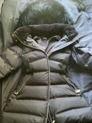 Svart pufferjacka från ZARA med fuskpäls - Snygg svart pufferjacka från ZARA med stor huva med fusk päls. Lång och varm och aldrig använt. Passar S-M