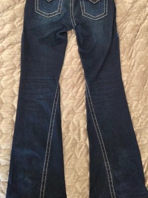True Religion Mid Rise Flare Jeans - Mörkblå jeans från True Religion med utsvängda ben och kontrastsömmar. Modellen är Mid Rise Flare och har dekorativa bakfickor med vita och blå sömmar. Tillverkade i denim med klassisk femficksdesign och knappgylf. Orginal pris 1200. SKRIV FØR KJØPING