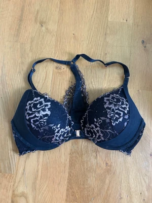 Svart Spets-Bh - Hunkemöller - Vacker svart bh med vadderade kupor och blommigt spetsmönster i ljusrosa. Axelbanden är justerbara och bh:n har en dekorativ spetsdetalj mellan kuporna samt framtillknäppning. Feminin och vacker. Använd fåtalet gånger och i fint skick. Inköpt på Hunkemöller. 