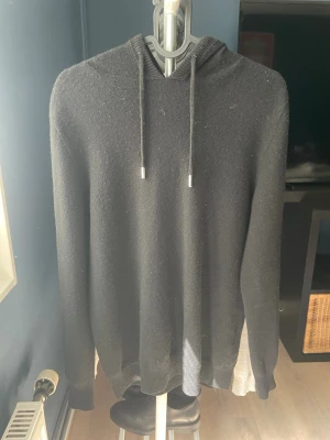Svart hoodie stickad - En stilren svart hoodie med huva och dragsko. Hoodien har långa ärmar och ribbade muddar vid ärmslut och nederkant. Perfekt för kalla dagar och enkel att matcha med olika outfits.