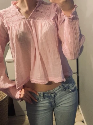 Ljusrosa blus med volangdetaljer - Söt ljusrosa blus i ett tunt, luftigt tyg med volangdetaljer vid axlar och halsringning. Blusen har lång ärm med resår vid ärmslut och en lös, flowy passform. Perfekt för en romantisk och avslappnad stil.