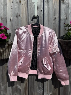 Rosa satin bomberjacka (XS) - Snygg rosa bomberjacka i glansigt satinmaterial med ribbade muddar och krage. Jackan har två fickor framtill med knappstängning och svart quiltat foder. Dragkedja framtill och klassisk bomberjacke-look.