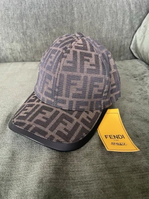 Fendi keps med FF-mönster - Säljer en snygg keps från Fendi med det klassiska FF-logomönstret i brunt och svart. Kepsen har böjd skärm med svart kant och justerbart band bak i läder. Tillverkad i tyg med exklusiv känsla och detaljer i läder. Perfekt för dig som gillar lyxiga accessoarer.