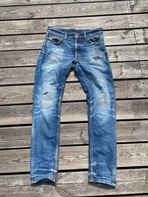 Blåa nudie jeans lean dean - Säljer ett par blå jeans med slitningar och raka ben. Jeansen har klassisk femficksdesign, ljus tvätt och detaljerade slitningar framtill och baktill för en trendig look. Perfekta för dig som gillar en avslappnad och cool stil. Modellen är lean Dean