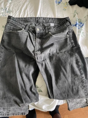 Grå denimjeans från H&M - Säljer ett par grå denimjeans från H&M med klassisk femficksdesign och knappgylf. Shortsen har en rak passform och är perfekta för en avslappnad stil. Tillverkade i bomull för en skön känsla hela dagen.