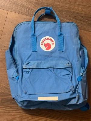 Blå Fjällräven Kånken ryggsäck - Klassisk blå Kånken ryggsäck från Fjällräven med ikonisk röd logga framtill. Ryggsäcken har ett stort huvudfack, en framficka med dragkedja och två sidofickor. Tillverkad i slitstarkt syntetmaterial med justerbara axelremmar och dubbla handtag upptill.