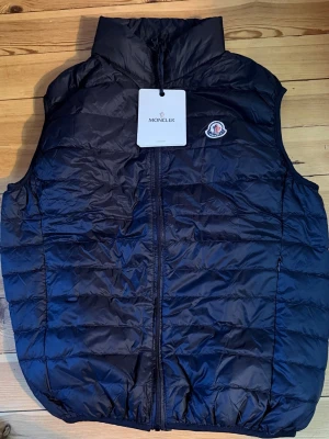 Stilren klassisk väst  -  Stilren Moncler-väst i klassiskt svart utförande.( ej äkta) Perfekt för både vardag och resa. Lätt, varm och enkel att matcha med resten av garderoben.   