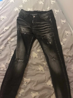 Svarta slitna jeans med skinny fit - Säljer ett par svarta skinny fit jeans från Dsquared  med slitningar och snyggt tvättad look. Jeansen har klassisk femficksmodell, knappgylf och läderpatch bak i midjan. Perfekta för en trendig streetstil.