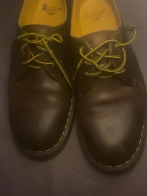 Bruna Dr. Martens loafers i läder - Säljer ett par bruna loafers från Dr. Martens i äkta läder med gula kontrastsömmar och matchande gula skosnören. Skorna har en robust sula med grovt mönster och klassisk Dr. Martens-design