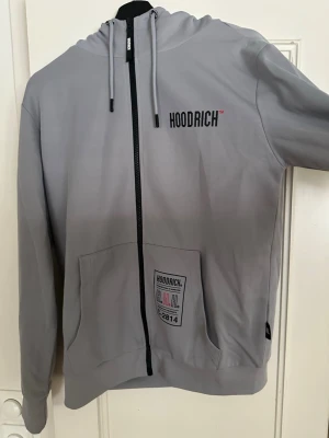Grå hoodie från Hoodrich med tryck - Snygg grå hoodie från Hoodrich med svart dragkedja, tryck både på bröstet, ryggen och ärmen. Huva med dragsko och coola street-inspirerade detaljer. Perfekt för dig som gillar streetwear och vill sticka ut.