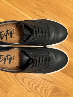 Svarta Eytys sneakers i läder - Stilrena svarta sneakers från Eytys i slätt läder med vita tjocka sulor och svarta skosnören. Klassisk låg modell med diskret lila hälflik och korkinnersula för extra komfort. Perfekta för dig som gillar minimalistisk och clean stil.