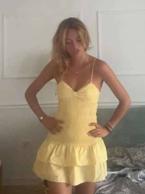 Zara Gul kortklänning med volang och rosett - Söt gul kortklänning från Zara med smala axelband, v-ringad front och volang nedtill. Klänningen har öppen rygg med en stor rosett som detalj. Perfekt för sommarens varma dagar och har en lätt och luftig känsla.