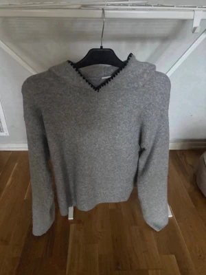 Grå stickad hoodie med svart kantdetalj - Mysig grå stickad hoodie med huva och snygg svart kantdetalj längs huvan. Tröjan har en enkel och stilren design med långa ärmar och mjukt material, perfekt för en avslappnad look.