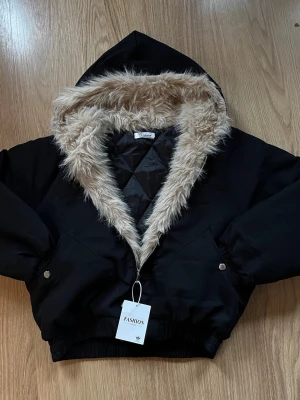 Svart pufferjacka med fuskpäls - Snygg svart pufferjacka med stor huva och beige fuskpäls runt kragen. 