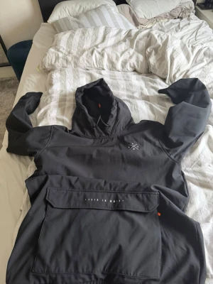 Svart dope skidjacka  - Svart dopejacka med stor magficka och huva, samt dragsko med orange detaljer. Liten vit logga på bröstet och elastiska muddar vid ärmslut och nederkant. Perfekt för skidor eller snowboardåkning 