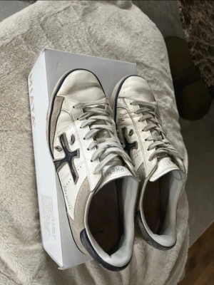 Premiata sneakers - Snygga vita sneakers från Premiata med mörkblå detaljer och logga på sidan. Skorna har klassisk låg profil, vita skosnören och ovandel i läder med inslag av mocka. Perfekta för dig som gillar stilrena och tidlösa sneakers.