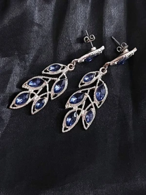 Silverfärgade örhängen med blå stenar - Snygga örhängen i silverfärgad metall med blå, glittrande stenar i bladformad design. Hängande modell som ger ett elegant intryck. Perfekta för att lyfta din stil med en touch av färg och glamour.