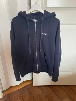 Marinblå hoodie från Dondup - Mörkblå hoodie med dragkedja från Dondup. Klassisk modell med huva, dragsnören och två stora fickor framtill. Liten vit logga på bröstet. 