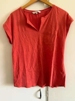 Röd kortärmad topp från Part Two - Snygg röd topp från Part Two med korta ärmar och liten bröstficka. Toppen har en avslappnad passform och en unik v-ringad detalj vid halsen. Perfekt att styla med jeans eller kjol för en cool look.