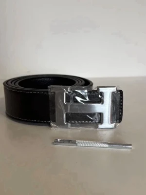 Svart Hermès bälte med silver H-spänne - Snyggt svart bälte från Hermès med klassiskt stort H-spänne i silverfärg. Bältet är gjort i skinn med vit kontrastsöm längs kanterna och har en stilren, tidlös design som passar till många olika outfits.