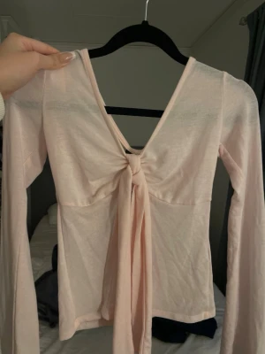 Ljusrosa långärmad topp Gina Tricot - Supersöt ljusrosa långärmad topp från Gina Tricot med knyt framtill och djup v-ringning. Tillverkad i mjukt och tunt material som känns skönt mot huden. Perfekt för att skapa en trendig och feminin look. (kolla ginas hemsida för fler bilder)
