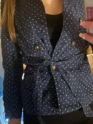 Mörkblå prickig trenchcoat med knytskärp - Snygg mörkblå trenchcoat med vita prickar och knytskärp i midjan. Jackan har dubbelknäppning med guldiga knappar och klassisk krage. Perfekt för dig som vill ha en trendig och unik look. Långärmad och i lätt tyg.