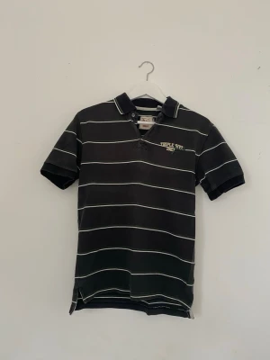 Vintage svart randig polotopp från Triple Five Soul - Cool svart polotopp från Triple Five Soul med vita och gröna ränder. Klassisk krage och korta ärmar, broderad logga på bröstet. Perfekt för dig som gillar streetwear och vill ha en avslappnad look. Y2K från 2000- talet