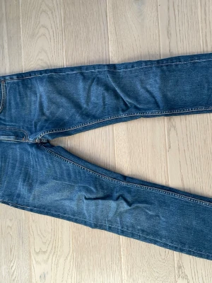 Blå jeans från Nudie Jeans, 29/32 - Säljer ett par klassiska blå jeans från Nudie Jeans i storlek 29/32. Jeansen har snygga kontrastsömmar och ikoniska broderade bakfickor. Perfekta för dig som gillar en tidlös och avslappnad stil.