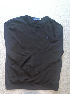 Svart sweatshirt från Polo Ralph Lauren - Svart sweatshirt från Polo Ralph Lauren med klassisk rund halsringning och broderad logotyp i blått på bröstet. Tröjan är tillverkad i mjukt bomullsmaterial och har ribbade muddar vid ärmslut och nederkant. Perfekt för en avslappnad och stilren look.