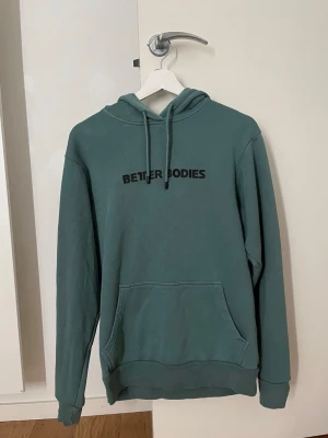 Grön hoodie från Better Bodies - Snygg grön hoodie från Better Bodies med svart tryck på bröstet. Hoodien har huva med dragsko och en stor magficka framtill. Perfekt för en avslappnad och sportig stil. Tillverkad i mjukt material för extra komfort.