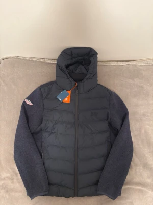 Superdry cardigan hoodie mörkblå - Snygg svart hoodie-cardigan från Superdry med vadderad framsida och stickade mörkgrå ärmar. Jackan har dragkedja, huva och Superdry-logga på ärmen. Perfekt för lager-på-lager och streetwear-stil. Självklart äkta och pris kan diskuteras