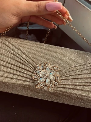 Glittrig guldig/Champagnefärgad kuvertväska/clutch/portmonä - Råkade köpa 2st av denna väskan. Har alltså inte använt denna. Perfekt storlek för att få plats med mobil och annat viktigt. Går att ha utan band eller med band. Snygg kuvertväska i glittrigt guldigt tyg med dekorativt kristallbroderi i mitten. Väskan har en elegant kedja i guldfärg. Perfekt för dig som vill sticka ut med en lyxig accessoar.
