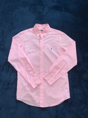 Rosa skjorta från Ralph Lauren - Klassisk rosa skjorta från Ralph Lauren med button-down krage och broderad blå logga på bröstet. Skjortan är i 100% bomull med stretch och har långa ärmar samt knappar framtill och vid ärmsluten. Perfekt för en clean och stilren look. 10/10 skick riktigt gött pris.