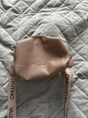 Beige axelväska från Valentino - Säljer en snygg beige axelväska från Valentino med justerbar axelrem där märkesnamnet står tryckt i svart. Väskan är i mjukt, strukturerat konstläder och har guldfärgade detaljer samt dragkedja upptill. Perfekt för dig som vill ha en stilren och trendig accessoar.