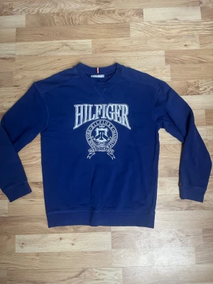 Blå sweatshirt från Tommy Hilfiger - Cool blå sweatshirt från Tommy Hilfiger med stort vitt tryck på bröstet och klassiskt Hilfiger-emblem. Tröjan har rund hals, ribbade muddar och är gjord i mjukt bomullsmaterial. Perfekt för en avslappnad och trendig look.
