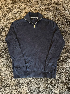 Mörkblå halvzip tröja Jack & Jones - Mörkblå stickad tröja från Jack & Jones med halvzip och hög krage. Tröjan har ribbade muddar vid ärmslut och nederkant, vilket ger en snygg och avslappnad look. Perfekt för lager på lager och enkel att matcha.