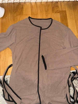 Brun kofta  - Mysig brun kofta med svarta kanter och knytband i midjan. Långa ärmar och två stora fickor framtill. Perfekt för chill dagar hemma. Materialet känns mjukt och bekvämt, troligen bomull eller bomullsblandning.