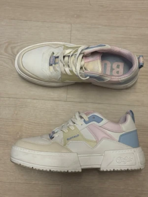 Buffalo sneakers i pastellfärger - Chunky sneakers från Buffalo i vitt med detaljer i pastellgul, ljusblå och ljusrosa. Skorna har en kraftig sula, snörning och logga på sidan och hälen. Tillverkade i syntetmaterial och textil för en modern och trendig look. Använt max 5 ggr 
