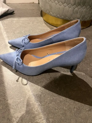 Ljusblå pumps med rosett från Zara - Snygga ljusblå pumps från Zara Trafaluc med spetsig tå och liten klack. Skorna har en söt rosettdetalj framtill och är klädda i ett tygmaterial. Perfekta för dig som vill ha en feminin och trendig look. Köpt på vinted men passar mig inte.