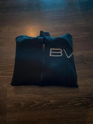 Svart hoodie från Bvaldi  - Snygg svart hoodie med dragkedja framtill och huva. På bröstet finns BV-logga i strass som ger en extra touch. Perfekt för dig som gillar stilrena plagg med lite bling. Mjuk och skön bomullsblandning. 