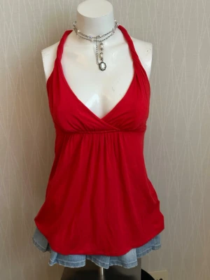 Röd ärmlös halterneck linnetopp - Snygg röd linnetopp med halterneck och omlott skärning framtill. Toppen är ärmlös och har en lätt utställd passform som ger en avslappnad look. Perfekt till jeans eller kjol för en trendig stil.