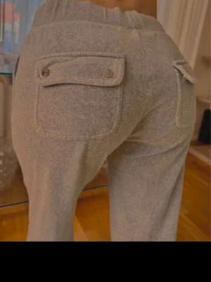 Juicy couture mjukis  - Säljer ett par grå mjukisbyxor med raka ben och två bakfickor med lock och knappar. Byxorna har bred resår i midjan och är gjorda i ett mjukt, bekvämt material som passar perfekt för chill dagar. Perfekt för dig som gillar en avslappnad och stilren look.