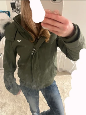 Grön vindjacka från Hollister - Snygg grön vindjacka från Hollister med avtagbar huva och fuskpäls. Jackan har dragkedja, knappar och ribbade muddar. Insidan är fodrad med mjuk teddy och loggan syns på bröstet. Perfekt för kyliga dagar och riktigt skön passform. den är mer mot s/ xs!
