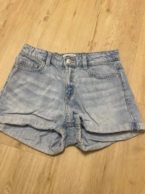 Ljusblå jeansshorts från Lindex - Snygga ljusblå jeansshorts från Lindex med klassisk femficksdesign, bälteshällor och uppvikta benslut. Perfekta för varma dagar och enkla att matcha med allt. Stängs med knapp och dragkedja framtill.