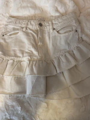 Beige volangkjol med jeanslook - Säljer en beige kortkjol med volanger och råa kanter. Kjolen har klassisk jeanslook med fickor fram och knappstängning. Perfekt för dig som gillar en avslappnad men trendig stil.