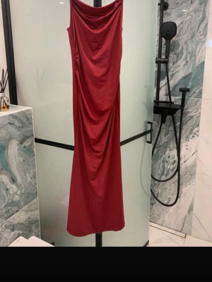 Röd lång klänning med drapering - Säljer en röd långklänning med snygg drapering framtill och breda axelband. Klänningen har en rak passform och är tillverkad i ett mjukt, stretchigt tyg som faller fint. Perfekt för dig som gillar en elegant och enkel look.